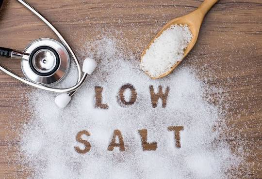 Stethoscope next to a wooden spoon and scattered salt spelling 'LOW SALT' on a wooden surface, conveying a message about a low-sodium diet.